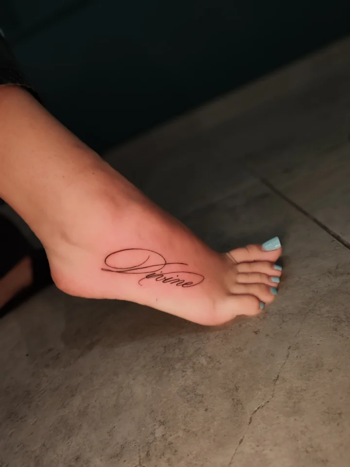 Tatuaggio sul collo del piede: la parola «Divine» in corsivo sottile, tratto continuo e senza ombreggiature.