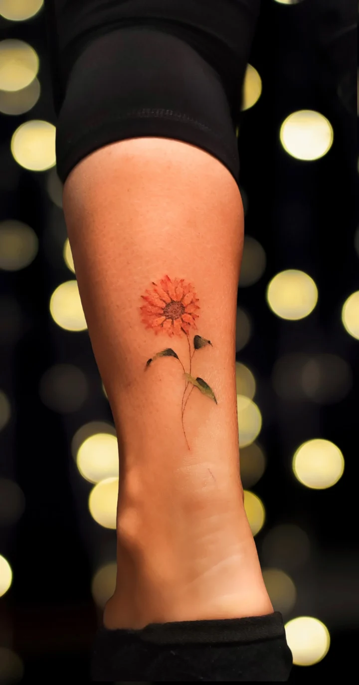 Tatuaggio sulla gamba: un girasole arancione dai petali sfumati, con foglie verdi e stelo sottile, in realismo a colori.