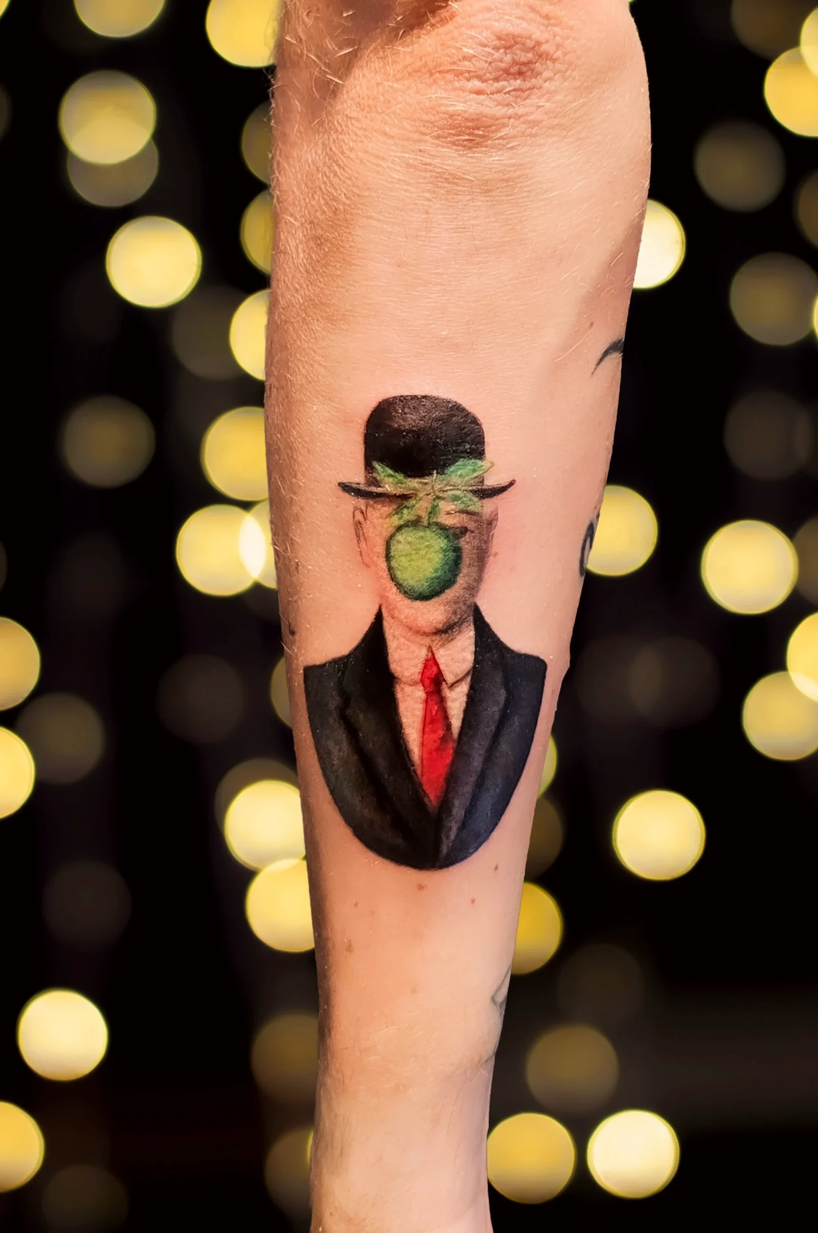 Tatuaggio sull'avambraccio: l'uomo con la bombetta e la mela verde di Magritte, in realismo a colori con la cravatta rossa.