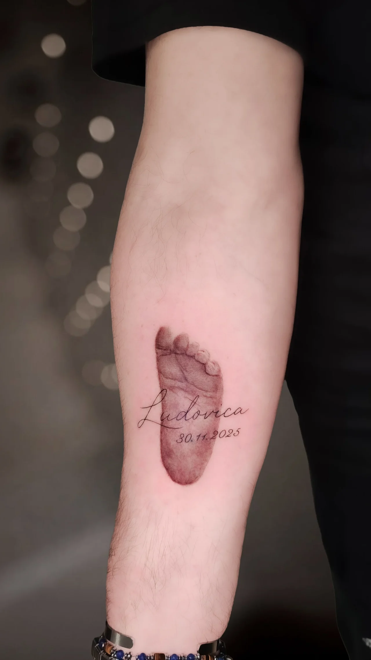 Tatuaggio sull'avambraccio: l'impronta di un piedino di neonato in realismo sfumato, con un nome in corsivo sottile e la data di nascita.