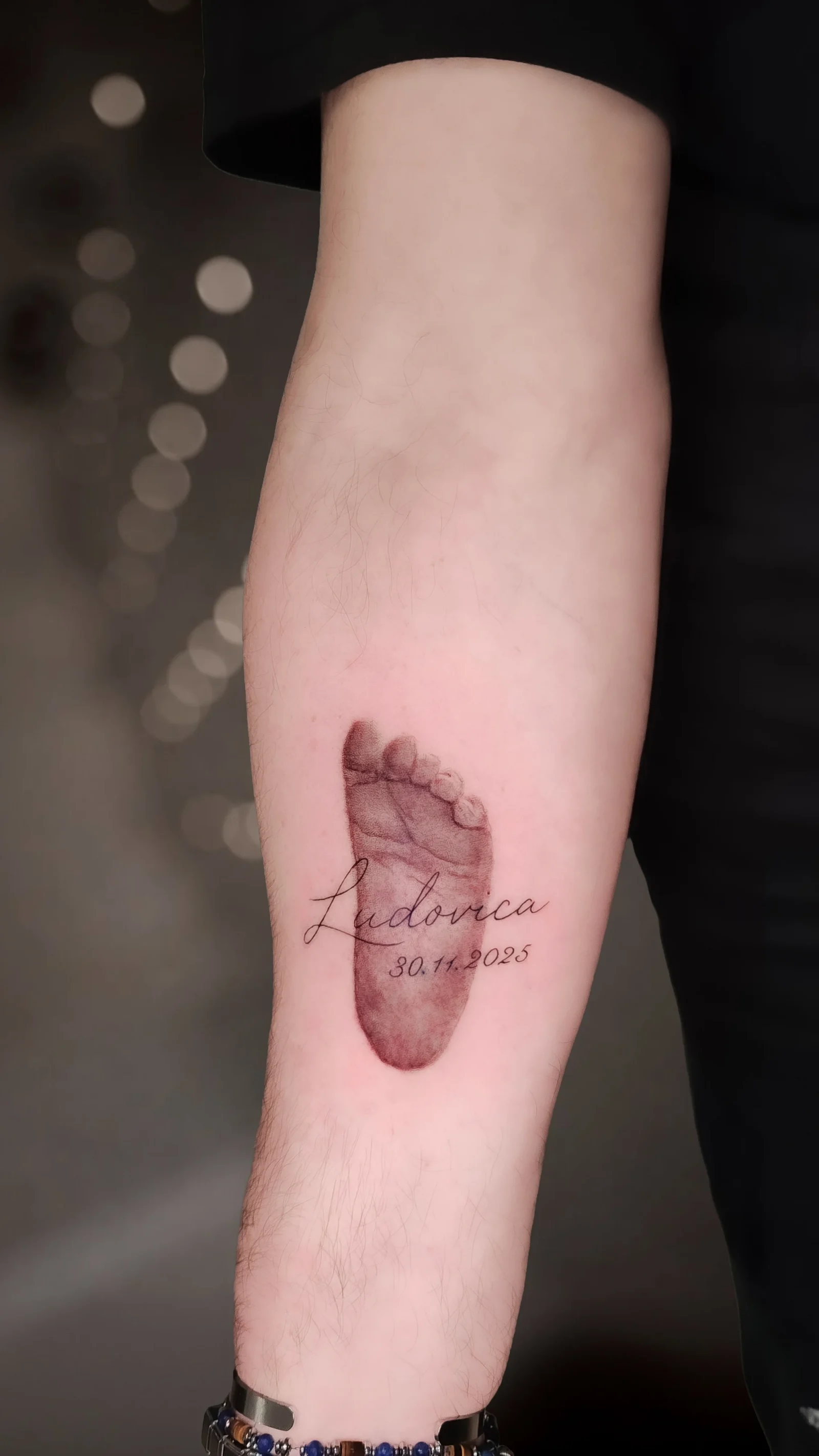 Tatuaggio sull'avambraccio: l'impronta di un piedino di neonato in realismo sfumato, con un nome in corsivo sottile e la data di nascita.