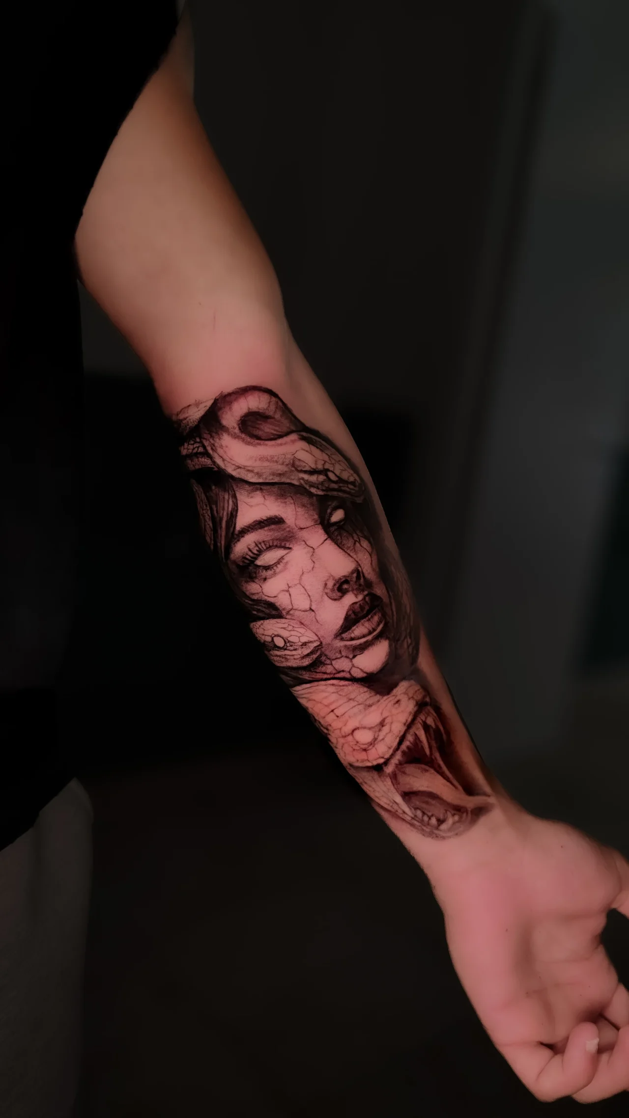 Tatuaggio sull'avambraccio: il volto di Medusa con gli occhi chiusi e la pelle screpolata, serpenti tra i capelli, in nero e grigio.