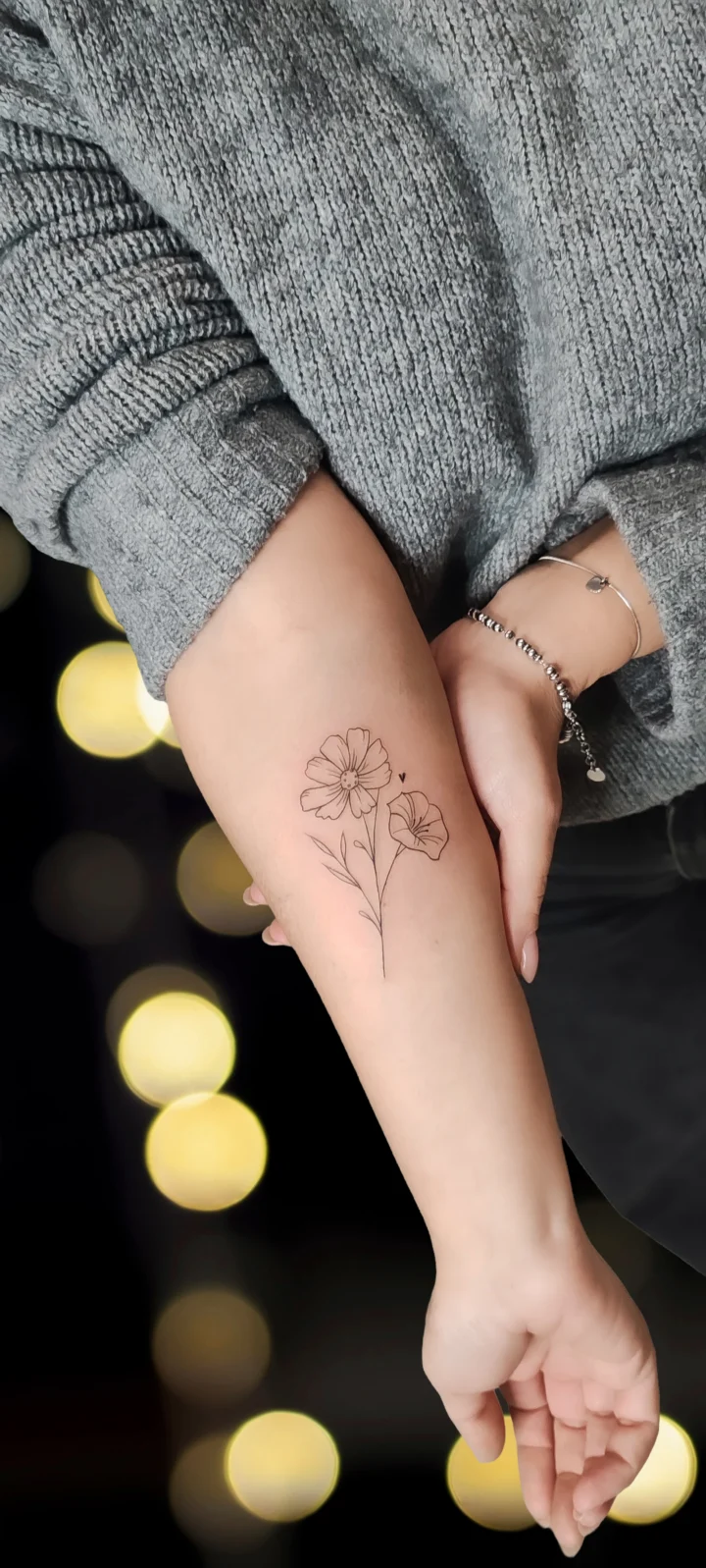 Tatuaggio sull'avambraccio: due fiori con stelo e foglie, disegnati a linea sottile continua e senza campiture.