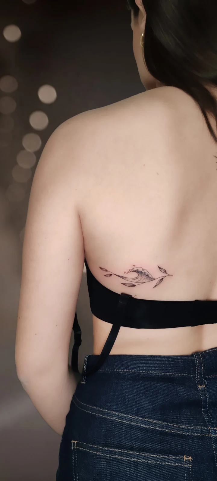 Tatuaggio sulla schiena: un'onda che si infrange tra piccole foglie, a linea sottile con una puntinatura leggera.