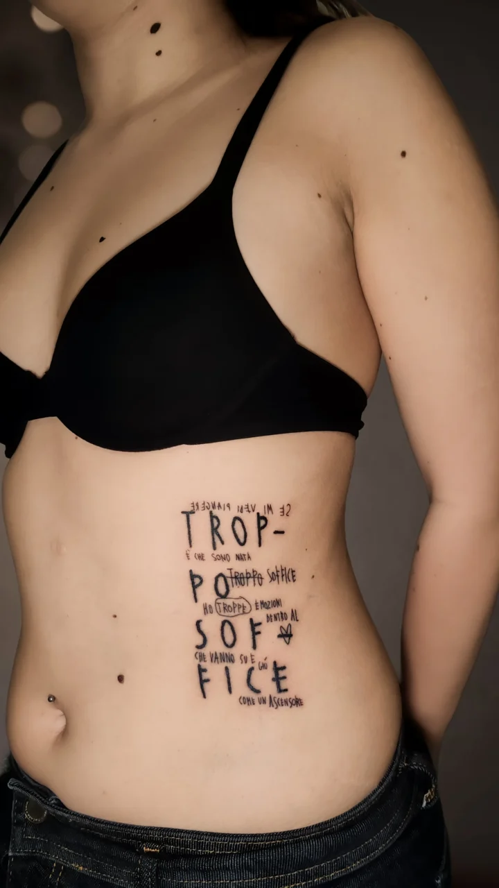 Tatuaggio sul fianco: la scritta «troppo soffice» in maiuscole disegnate a mano, con frasi più piccole intrecciate tra le righe.